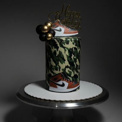 A custom cylindrical cake decorated with a green and beige camouflage pattern and featuring Nike sneakers near the base, topped with a gold 'Happy Birthday' script topper and gold beads, displayed on a silver cake stand against a dark background.
